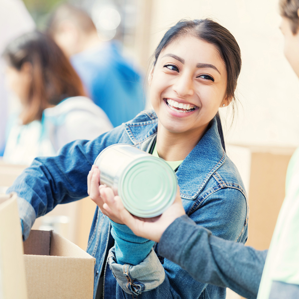 Confident Hispanic young woman accepts canned food items from people in her community during a food drive.